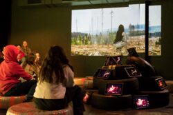 Teilnehmende sitzen auf Sitzkissen und schauen eine Projektion an. Ipads zeigen verschiedene Bilder und sind aufgetürmt