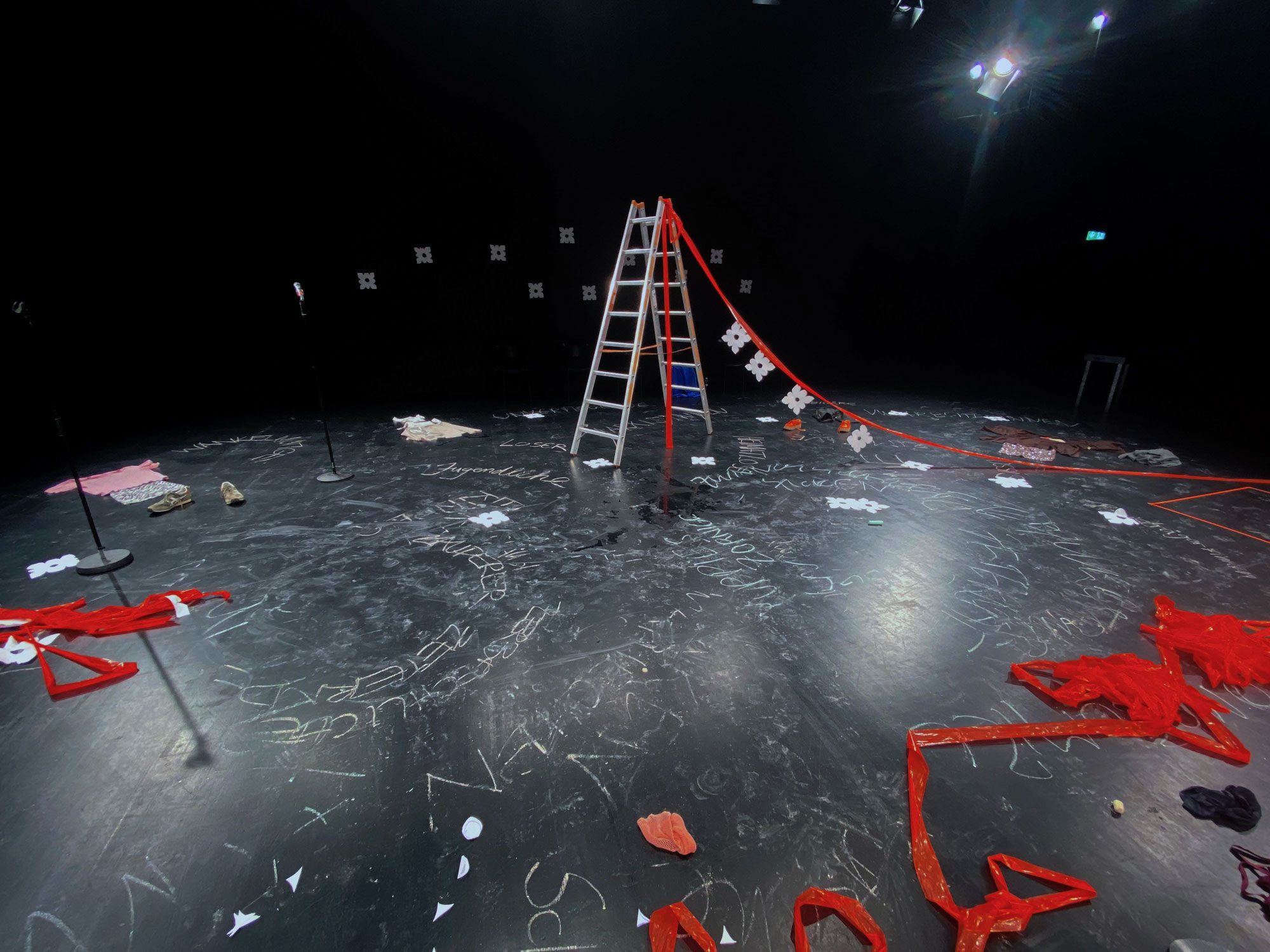 Verlassene Bühne übersät mit verschiedenen Dingen, Blättern Papier, mit Kreide sind Wörter auf den Boden geschrieben, eine Leiter steht in der Mitte